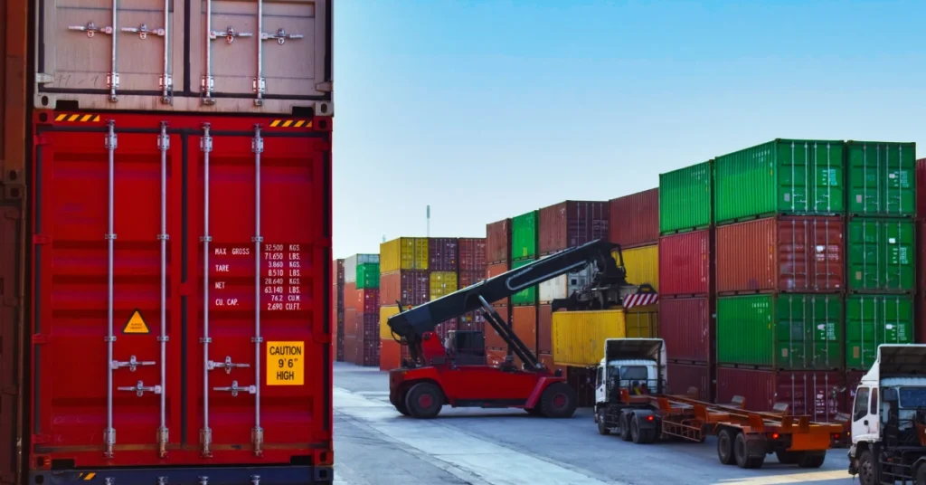 Red shipping container beside stacking truck in busy freight yard with colorful cargo containers after Amazon custom clearance