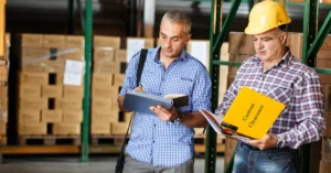 Two warehouse workers, one wearing a hard hat, review custom clearance documents amidst stacked boxes.