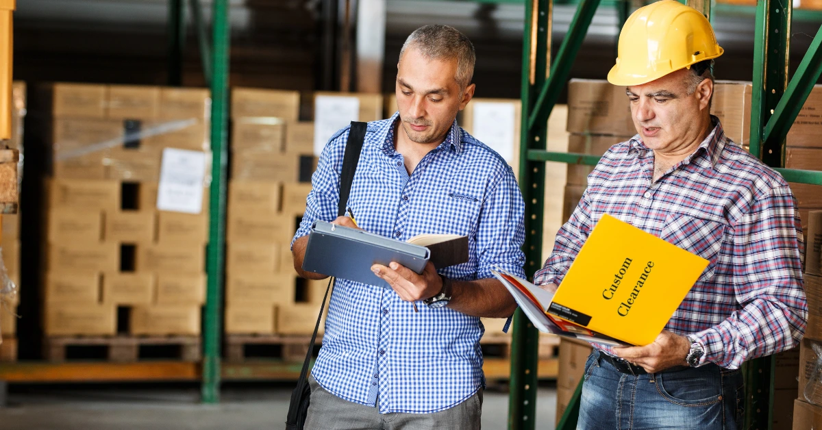 Two warehouse workers, one wearing a hard hat, review custom clearance in China documents amidst stacked boxes.