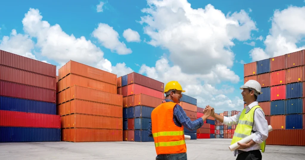Two workers in safety vests and hard hats shaking hands at shipping container yard, symbolizing successful product sourcing from China with colorful containers and blue sky in background