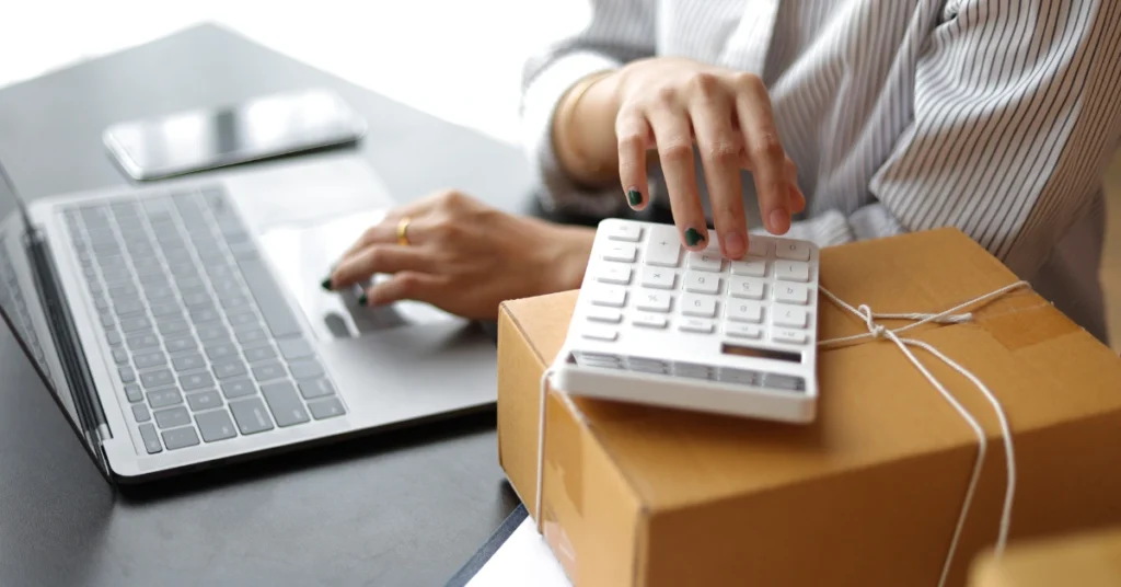 A person uses a calculator on top of a cardboard box while working on a laptop at a desk, likely calculating FBA shipping costs.