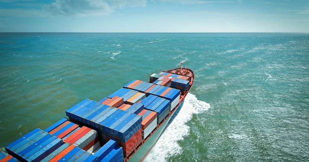 Cargo ship sailing on a vast ocean under a partly cloudy sky. Colorful shipping containers are stacked on the deck.